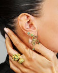 Close-up of a person wearing gold and green earrings with a hand holding more earrings.