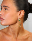 Close-up of a woman wearing gold earrings with green gemstones against a neutral background