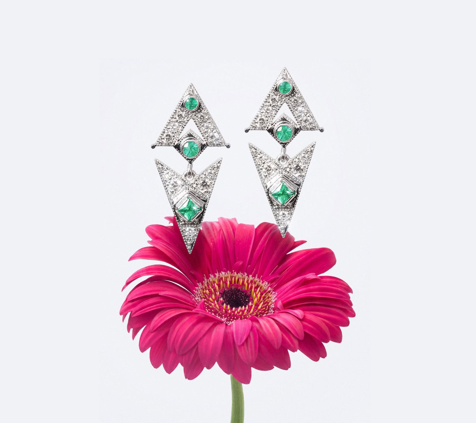 Diamond earrings with green gemstones on a pink flower against a light gray background