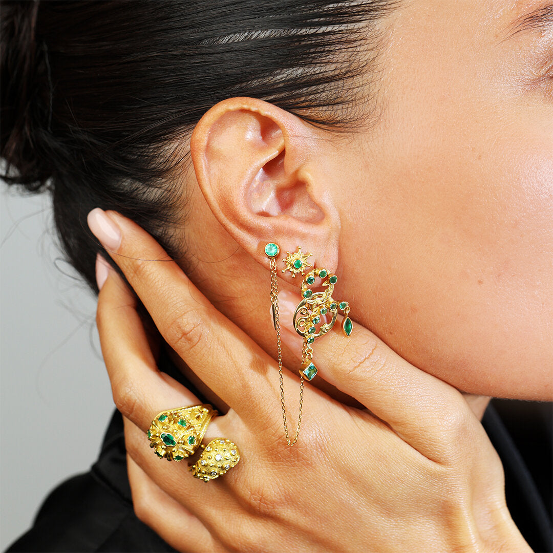 Close-up of a person wearing gold and green earrings with a hand holding more earrings.